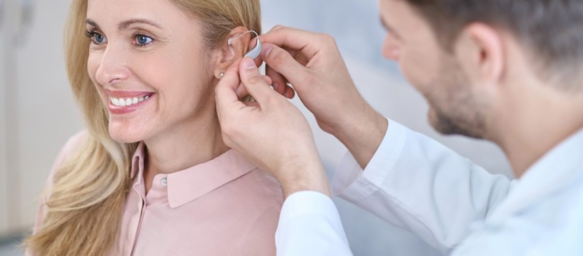 Fitting. Man in a white coat putting hearing aid on ear of smiling middle-aged woman with long blond hair sitting in medical office