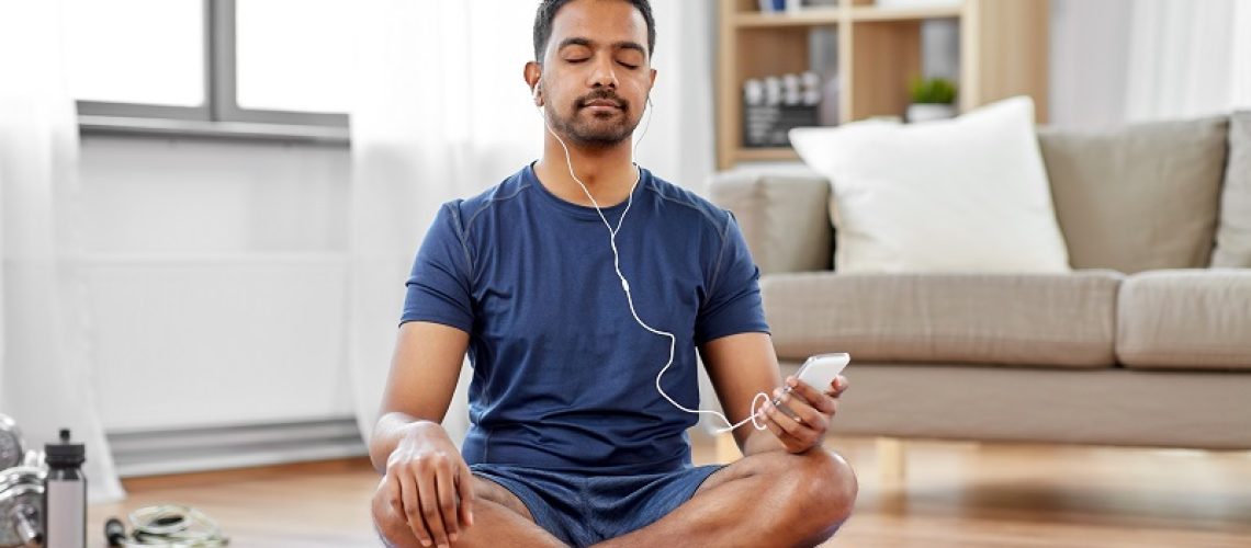 sport, technology and healthy lifestyle concept - indian man in earphones listening to music on smartphone at home