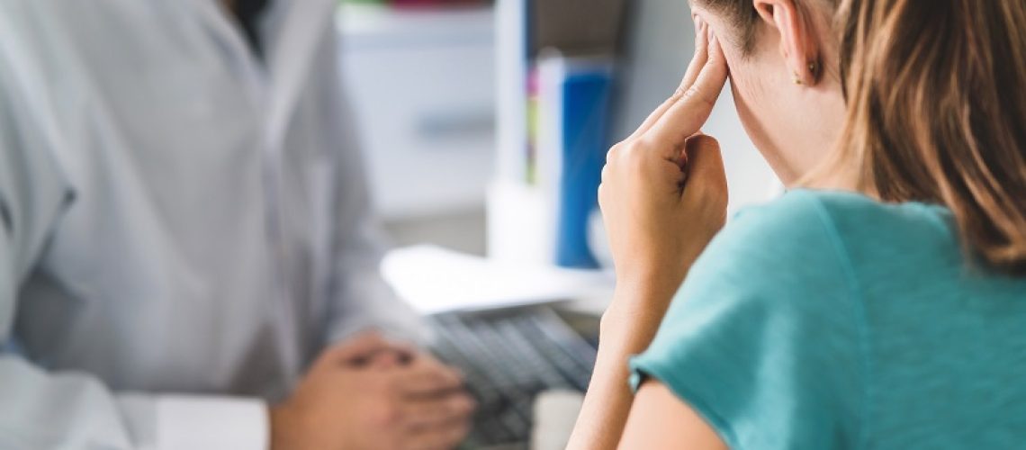 Woman suffering from bad headache or migraine. Appointment with doctor in office room. Sick and unwell lady with stress, trauma or burnout. Young patient in pain holding head with hands. Medical visit