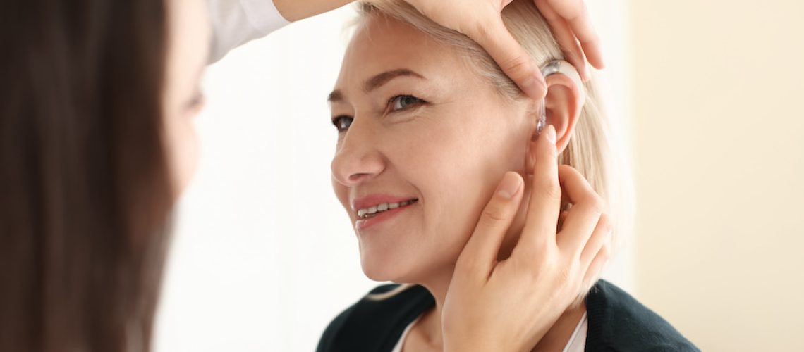 Otolaryngologist putting hearing aid in woman's ear on light background