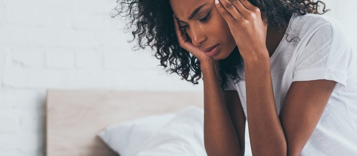 pretty african american woman holding hands near head while suffering from headache in bedroom